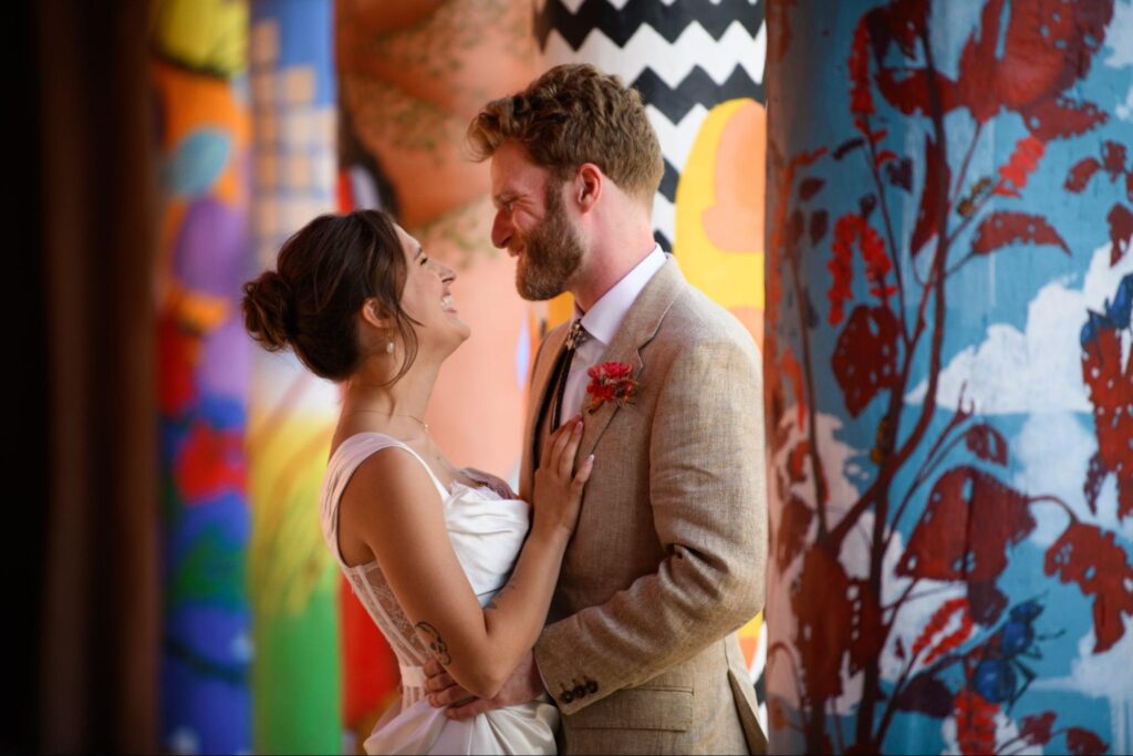 a bride and groom sharing a laugh while standing close together against a backdrop of brightly painted, artistic columns. showcasing how you can personalize your wedding and even your wedding photography.