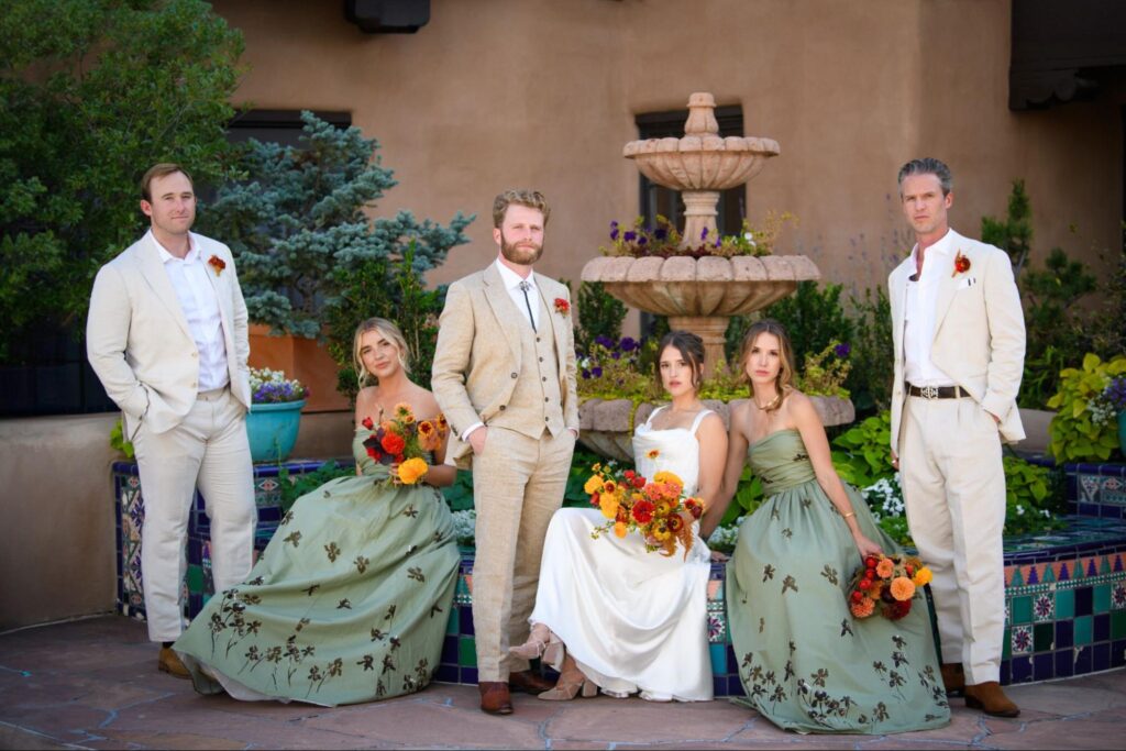 a wedding party posed by a Southwestern-style fountain, surrounded by greenery and colorful pottery. The groom wears a light beige suit, while the bride in a simple white gown holds a vibrant bouquet. Bridesmaids in sage-green dresses with floral embroidery and groomsmen in beige suits.