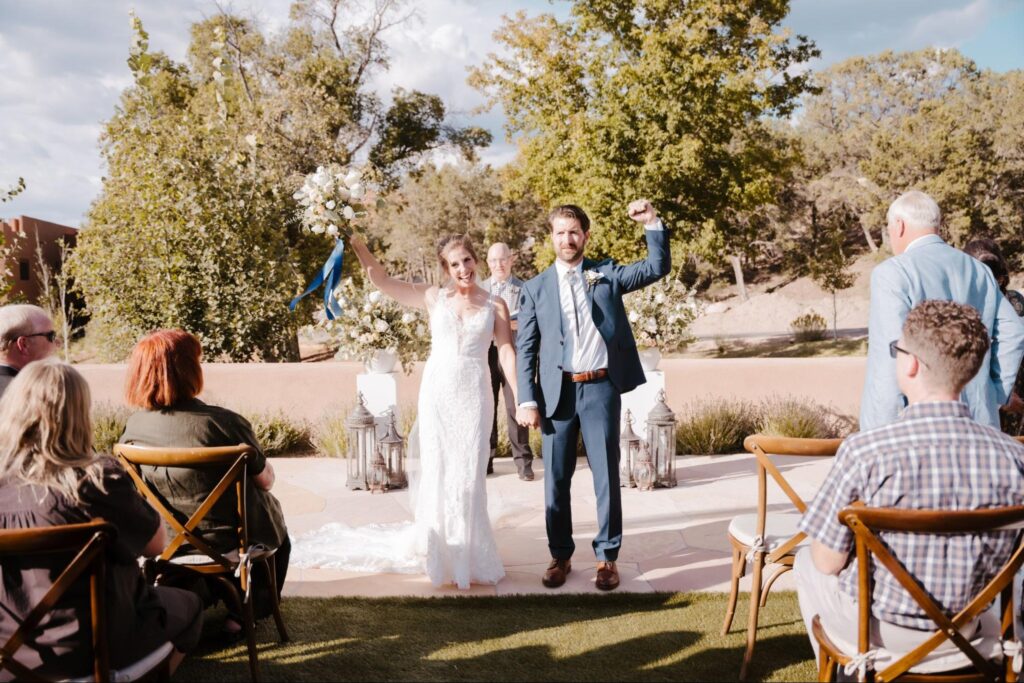 Happy couple celebrating their wedding at a Santa Fe wedding venue, sharing a joyful moment with their guests at an outdoor ceremony. The bride's holding her bouquet up, and the groom's raising his fist, all against a backdrop of trees and blue sky, creating a relaxed and beautiful vibe.