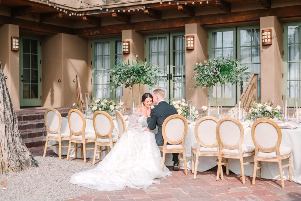 Elegant outdoor wedding setup at a Santa Fe wedding venue, featuring a beautifully decorated table with floral arrangements and greenery. The bride and groom share a sweet moment at the head of the table, surrounded by stylish chairs and warm adobe architecture.