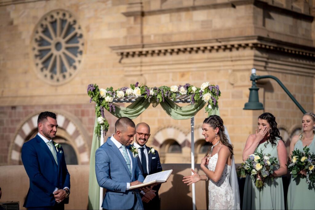 Outdoor wedding ceremony at a Santa Fe wedding venue, with the couple sharing a heartfelt moment under a beautiful floral arch. The bride and groom are surrounded by their wedding party, with a historic building as the backdrop.