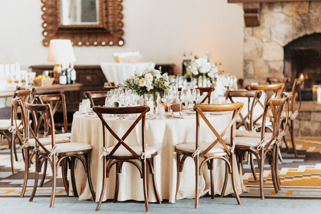 Charming indoor reception setup at a Santa Fe wedding venue, featuring a round table elegantly decorated with floral centerpieces, glassware, and candles. A wooden chairs and cozy ambiance, with a stone fireplace in the background.