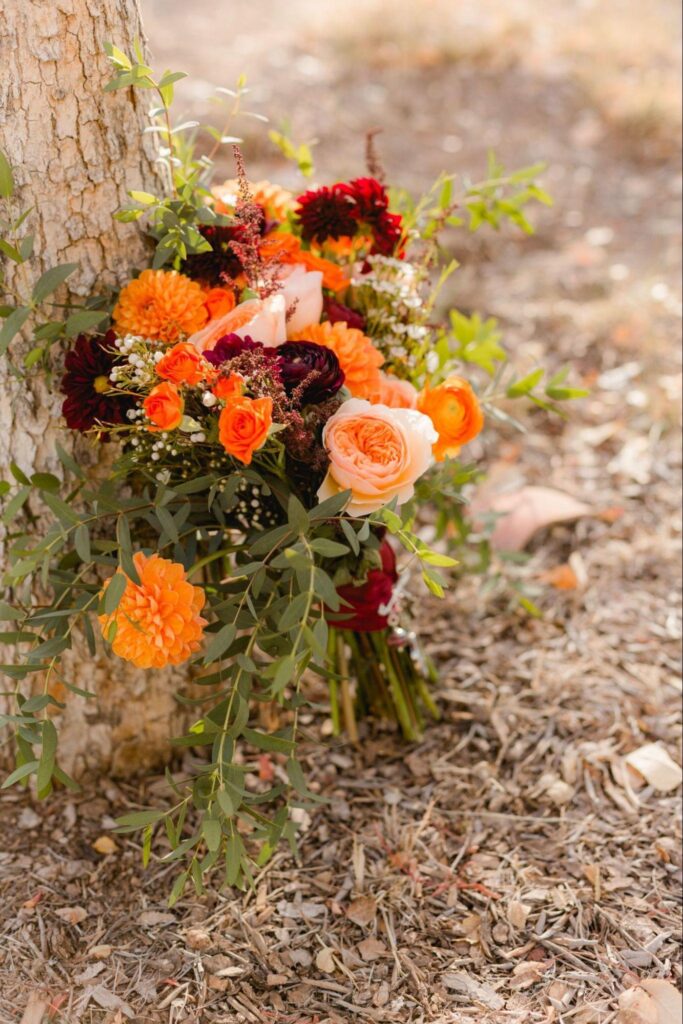 This photo showcases a vibrant wedding bouquet with orange, peach, and deep red flowers, beautifully arranged against a natural outdoor setting.