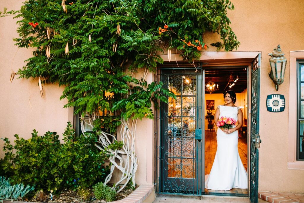 Bride in a white dress standing in a doorway with greenery and flowers around, adobe walls, and an iron gate.