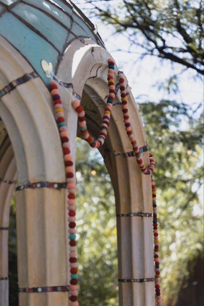 A close-up of a decorated arch with multicolored beads draped over its light stone structure, set against a background of trees.