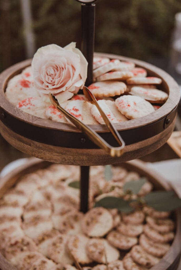 A tiered wooden tray displaying biscochitos, traditional New Mexican cookies, with a pink rose and serving tongs on top.