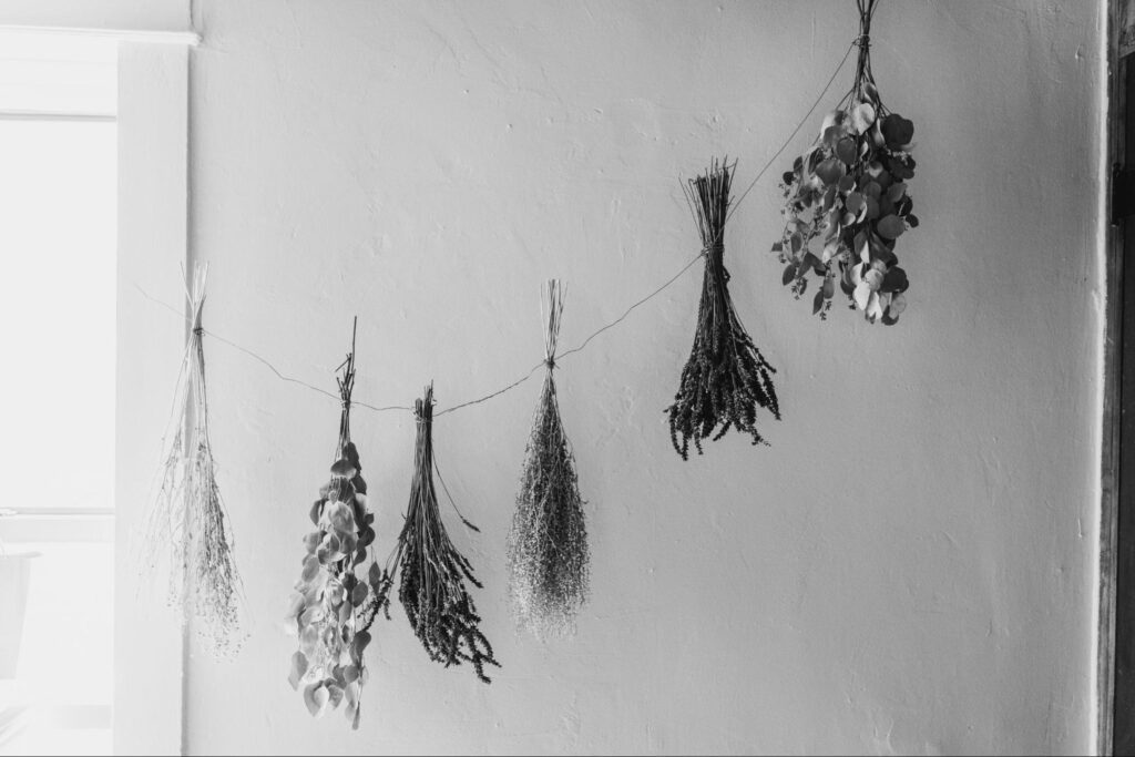 Black-and-white photo of dried flowers and greenery hanging on a string against a plain white wall.