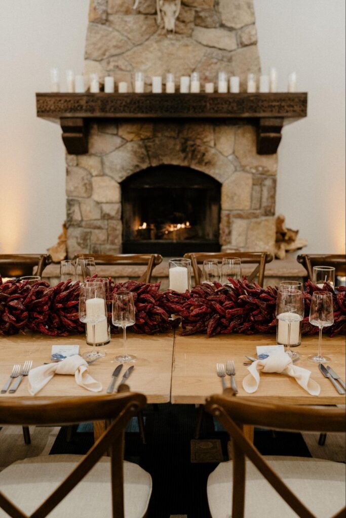 A stone fireplace with lit candles on the mantel, and a wooden dining table set with a centerpiece of dried red chiles and candles.