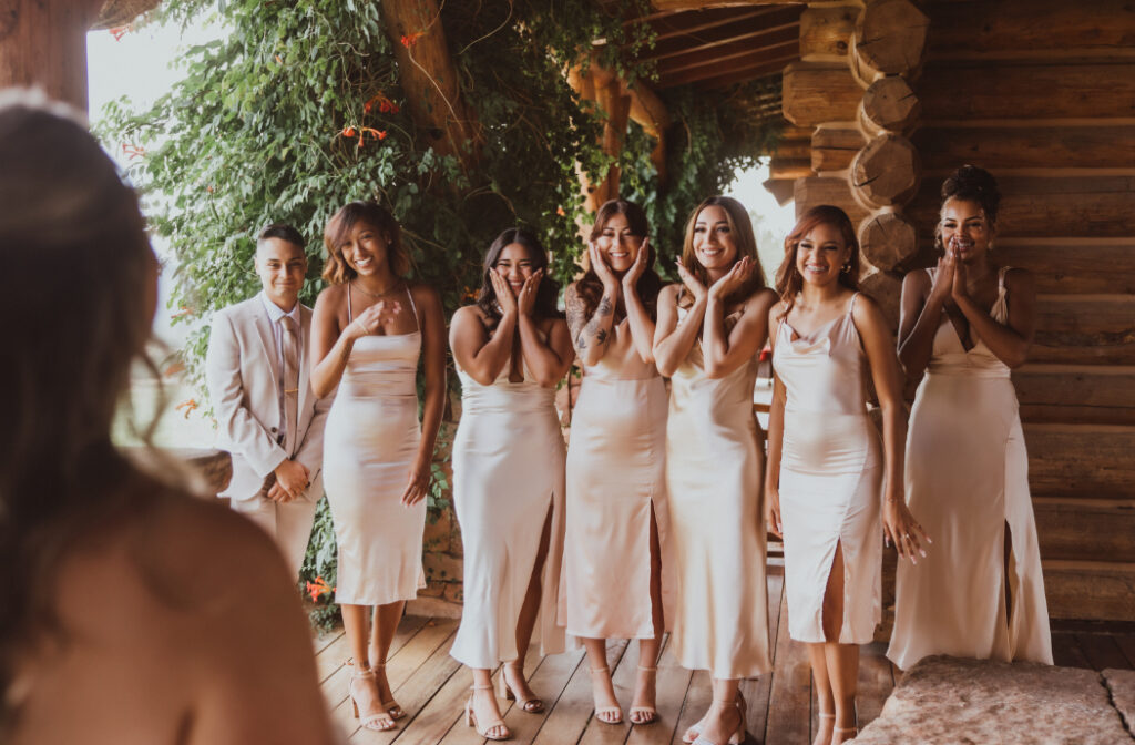 A group of bridesmaids in champagne satin dresses reacts with joy and surprise as they see the bride for the first time on a rustic wooden porch.
