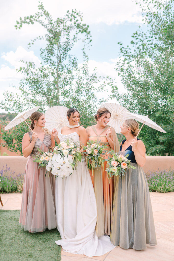 A bride in a white gown stands with her bridesmaids, holding white parasols and bouquets, against a lush outdoor Santa Fe wedding venue backdrop.