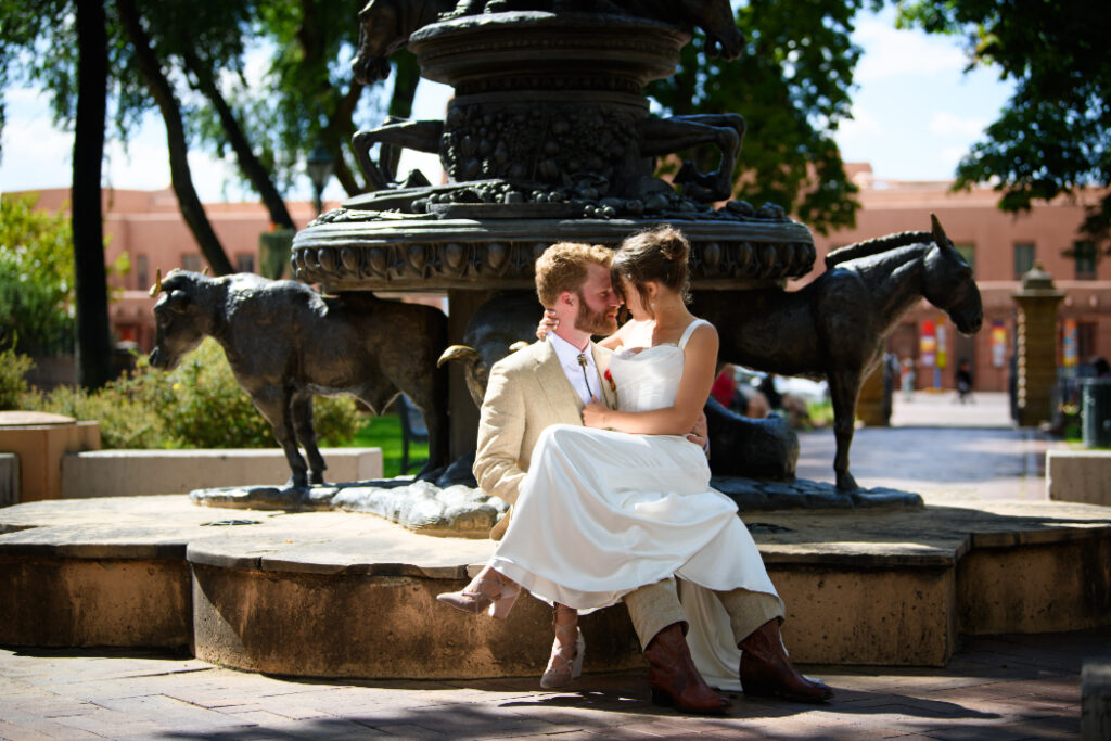 A newlywed couple sits on the edge of a bronze fountain featuring animal sculptures in a Santa Fe plaza. The groom, in a tan suit, and the bride, in a white dress with cowboy boots, embrace and smile at each other.