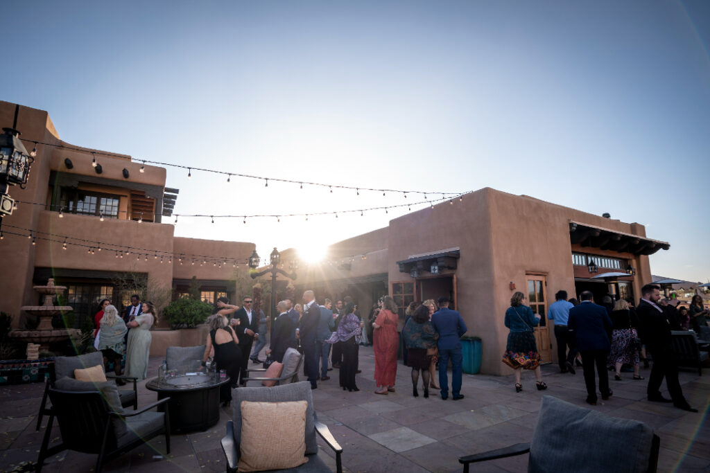 A wedding cocktail hour takes place on a rooftop terrace as the sun sets. Guests in formal and semi-formal attire mingle, while some sit around lounge seating. 