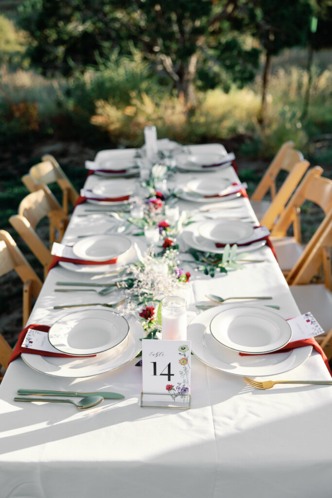 A beautifully set outdoor reception table features white linens, gold-rimmed plates, and red napkins. A floral centerpiece and a table number card add a polished touch under natural sunlight.