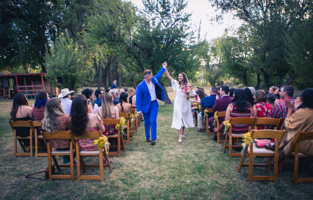 A joyful couple walks down the aisle after their outdoor ceremony, surrounded by seated guests in a lush, green setting. One wears a bold blue suit, and the bride carries a colorful bouquet.