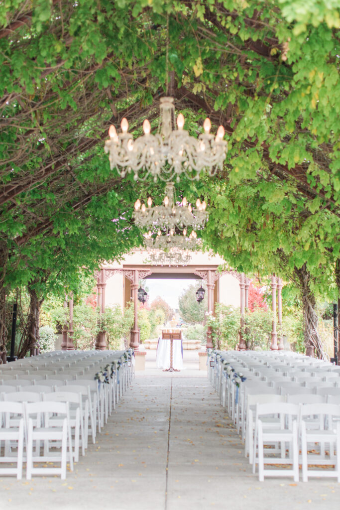 a wedding venue with a vine-covered archway, chandeliers, and white folding chairs set up for a ceremony.