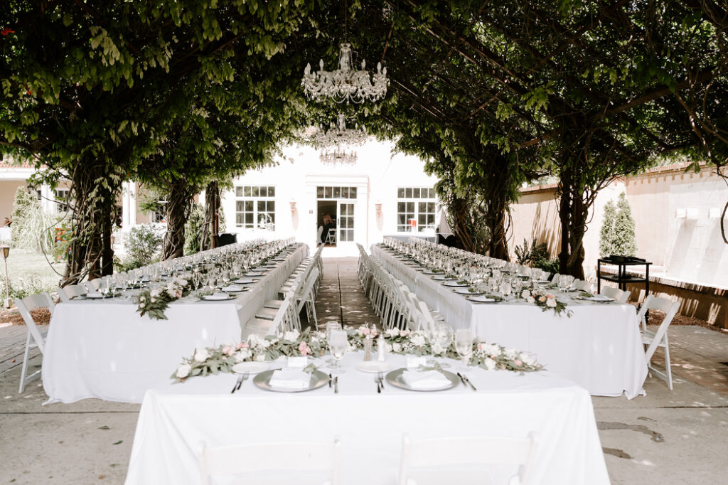 Long banquet-style outdoor reception setup featuring white table linens, floral centerpieces, and a symmetrical arrangement of tables beneath the chandeliers.