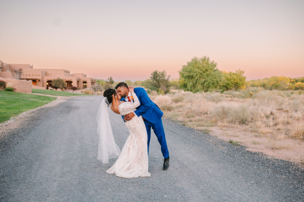 couple on a rural road at sunset. The groom, in a bold blue suit, dips the bride, who wears a long-sleeved lace wedding dress.