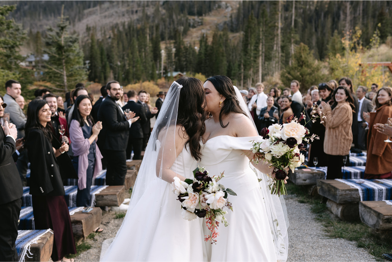 Brides share kiss at end of aisle. 