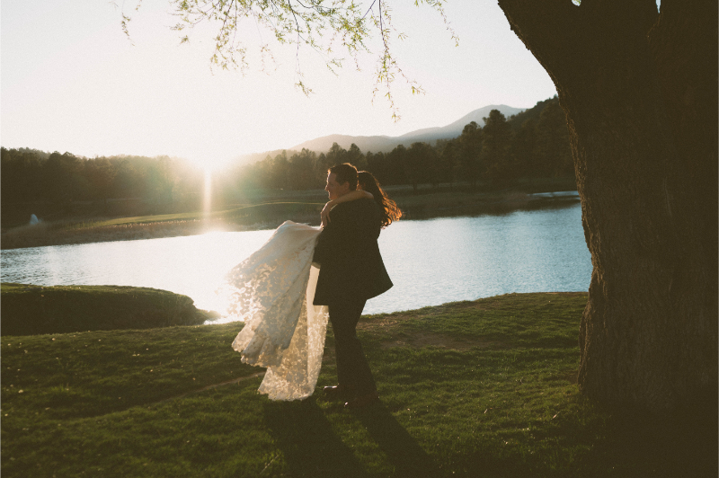 Groom twirls bride around in front of lake backdrop.