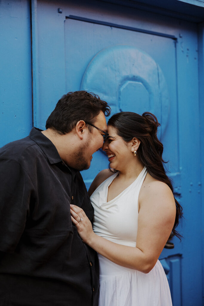 Couple stands with their heads together in front of blue door after proposal.