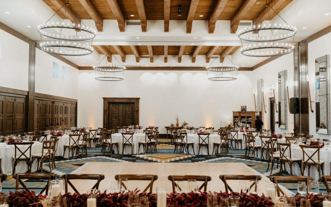 Elegant indoor reception area at one of the Santa Fe wedding venues, featuring a spacious room with wooden beams, large circular chandeliers, and round tables set with white linens. The decor includes rustic wooden chairs, candles, and floral arrangements.