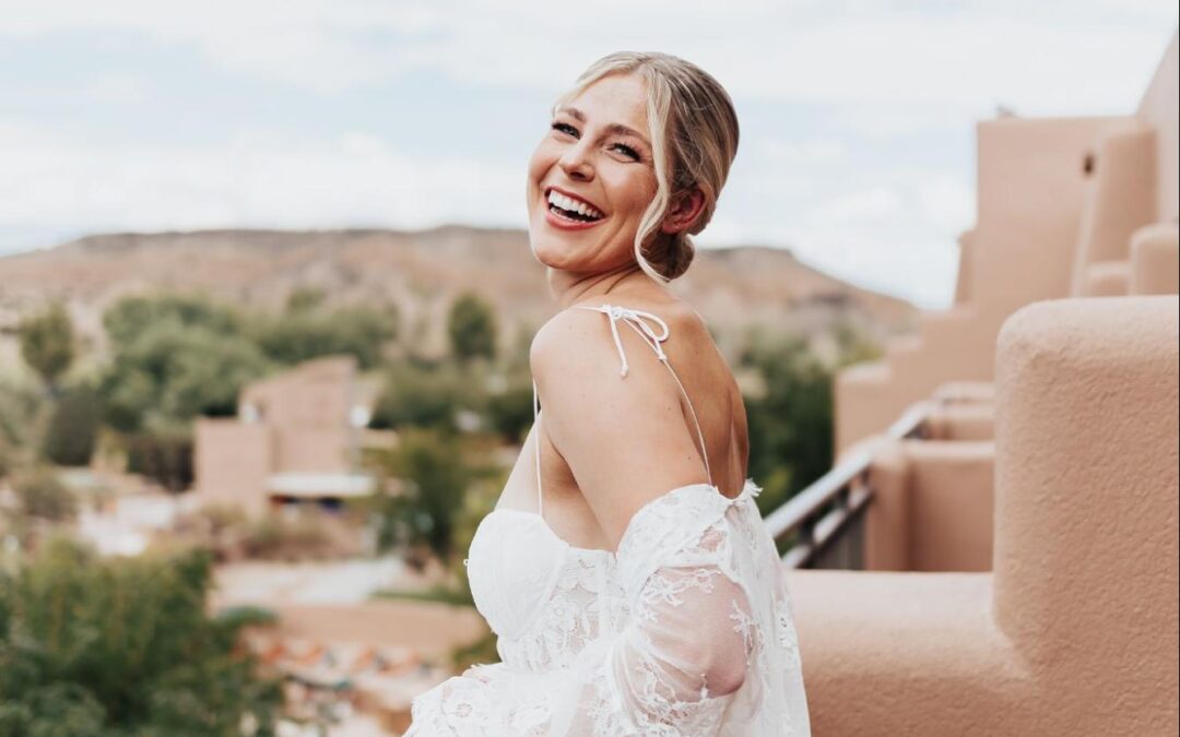 Bride laughing on a balcony with blue skies and adobe buildings in the background, wearing a lace off-the-shoulder gown. | outdoor wedding venues in albuquerque