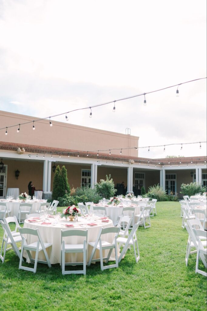 Outdoor reception setup with round tables, white chairs, and string lights on a grassy lawn near adobe-style buildings | outdoor wedding venues in albuquerque