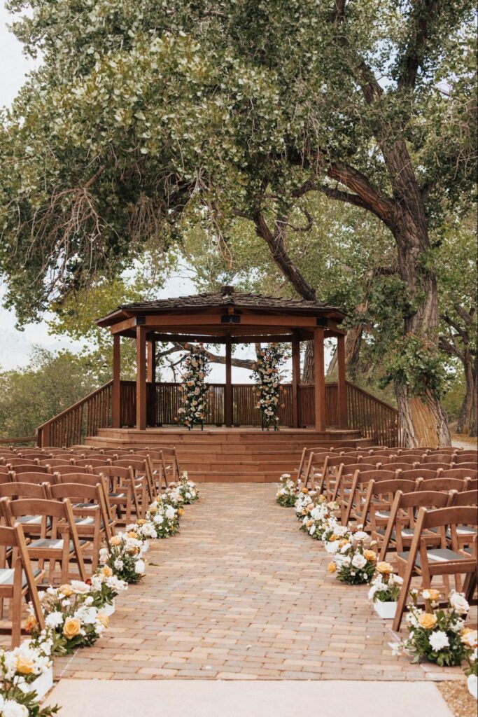 Ceremony site with a wooden gazebo, tree-lined backdrop, and flower-lined aisle featuring brown chairs on a brick pathway | outdoor wedding venues in albuquerque