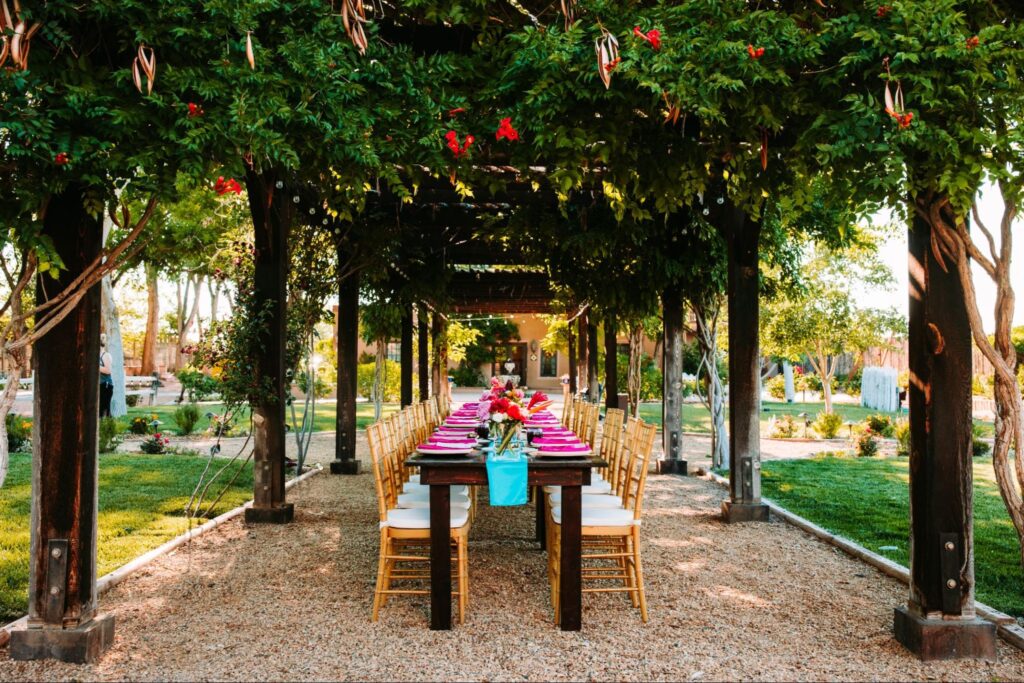 Long table setup under a shaded pergola with greenery and bright floral decor, surrounded by a lush garden.