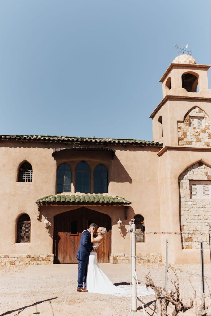 Couple kissing in front of a rustic adobe-style building with a tiled roof and blue skies above.