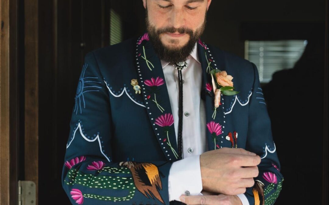 A groom adjusts his cufflinks while wearing a dark teal suit with colorful embroidery featuring cacti, flowers, and skull motifs.