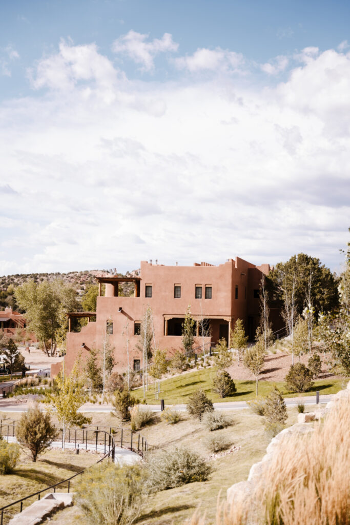 A Southwestern adobe-style building with earth-toned stucco, surrounded by greenery and rolling hills under a partly cloudy sky.