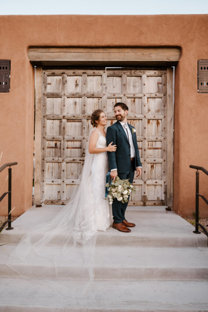 A bride and groom smile in front of a rustic wooden gate with adobe walls.