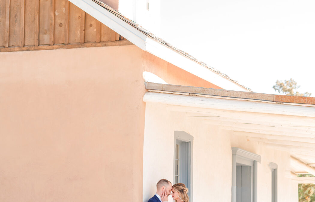 A bride and groom embrace beside the historic chapel at Bishop's Lodge Santa Fe, NM, with soft adobe tones and blue accents.