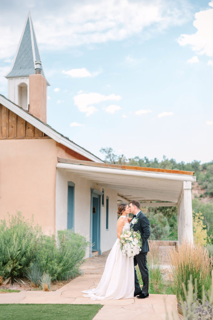 A couple kisses in front of the chapel at Bishop's Lodge Santa Fe, NM, surrounded by lush greenery.