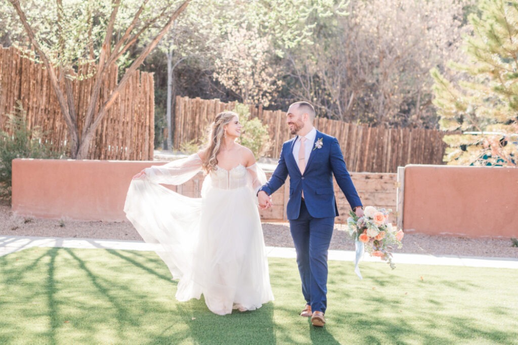 A bride and groom walk hand in hand on a lawn at Bishop's Lodge Santa Fe, NM, with wooden fencing and trees in the background.