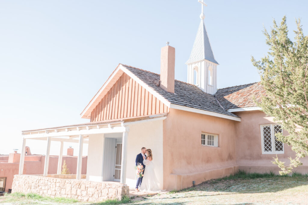 A bride and groom embrace outside the historic chapel at Bishop's Lodge Santa Fe, NM, with adobe walls and a white steeple.