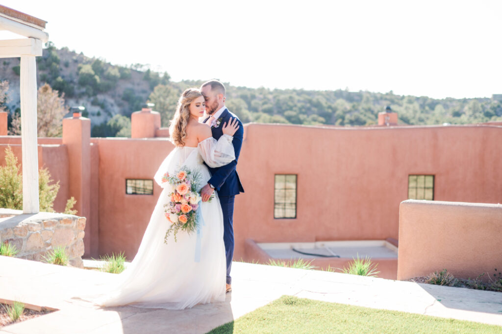 A bride and groom pose in front of an adobe building at Bishop's Lodge Santa Fe, NM, with rolling hills in the background.