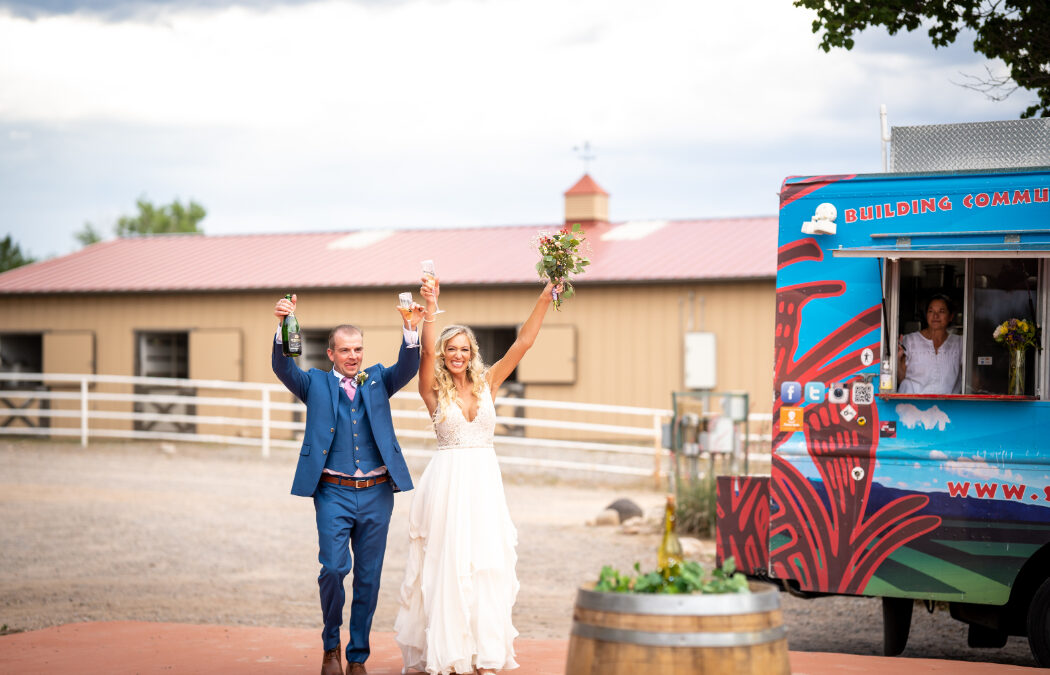 Bride and groom celebrate with champagne near a colorful food truck at an outdoor wedding venue.