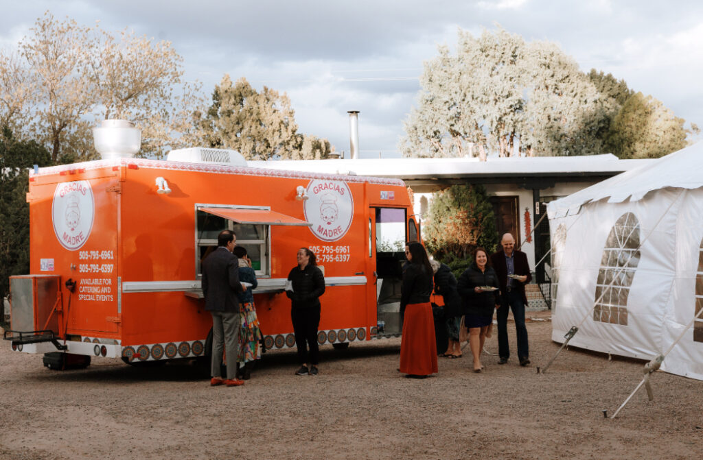 Guests order food from an orange food truck labeled "Gracias Madre" at an outdoor wedding reception.