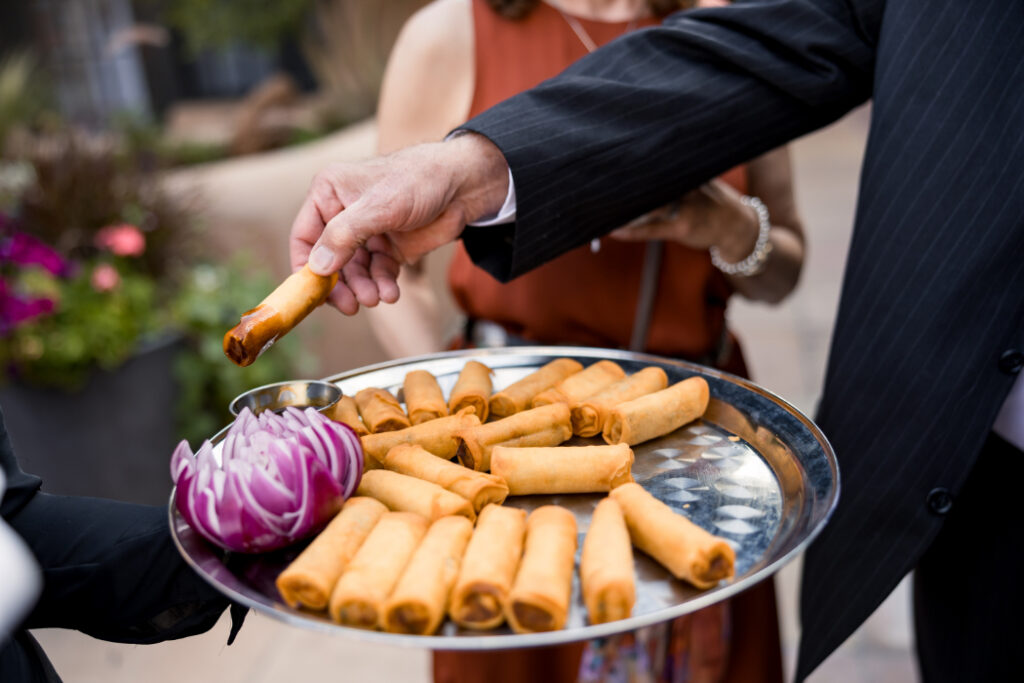 A guest reaches for a golden spring roll from a silver tray, garnished with carved onion. | catering in santa fe