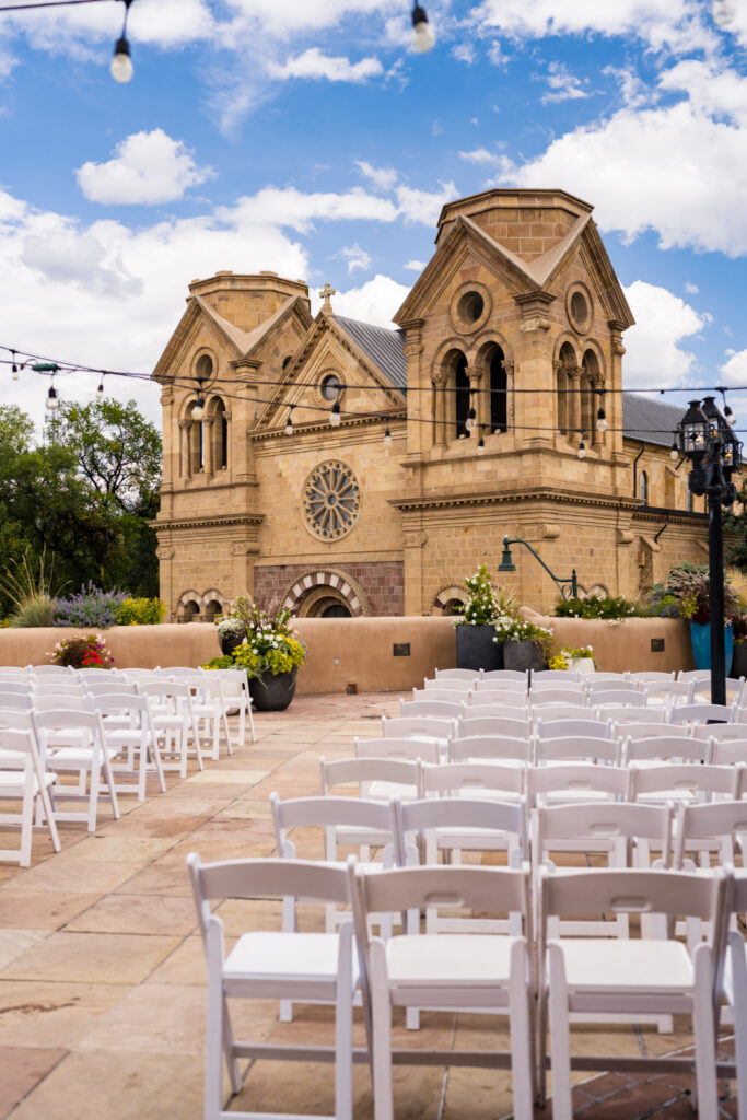 Outdoor wedding ceremony setup on a terrace with white chairs facing the Cathedral Basilica of St. Francis of Assisi in Santa Fe. String lights hang above, and potted flowers decorate the space.