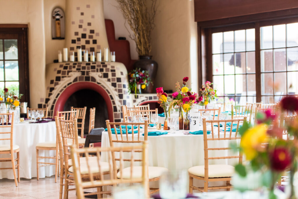 Reception space with round tables, colorful floral centerpieces, and teal napkins. A large adobe fireplace with mosaic tile details and candles serves as the backdrop in a Southwestern-style venue.