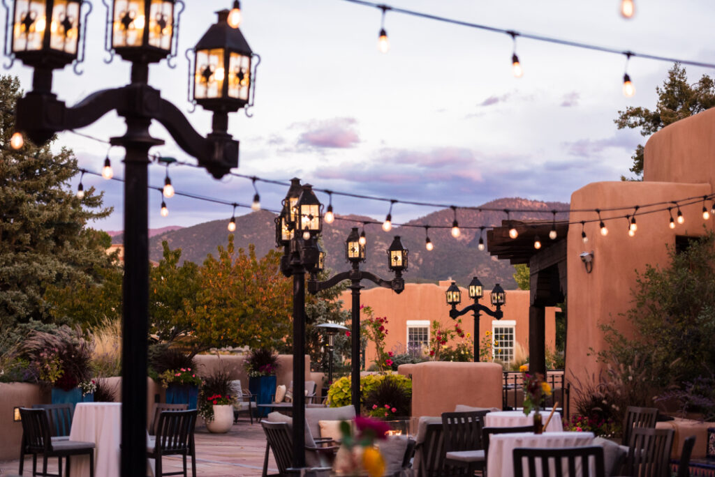 Rooftop terrace at sunset with adobe architecture, string lights, and vintage-style lanterns. Tables with floral arrangements and mountain views create a warm ambiance for an outdoor event.