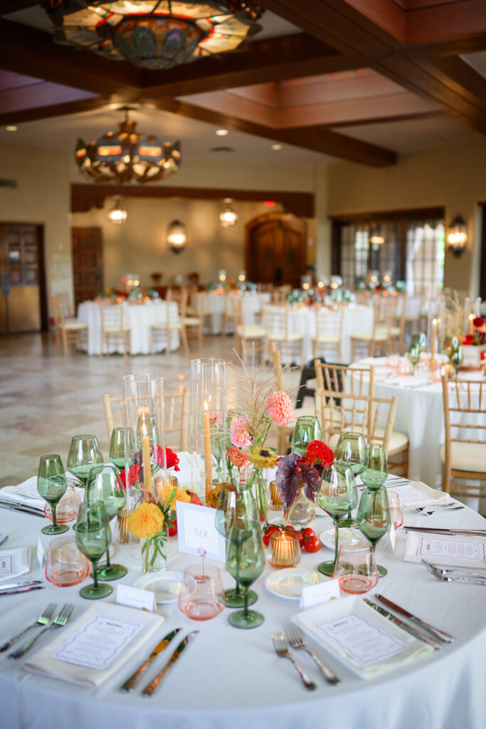 A wedding reception space is set with round tables covered in white linens. Each table features green glassware, colorful floral arrangements, and tall candles.