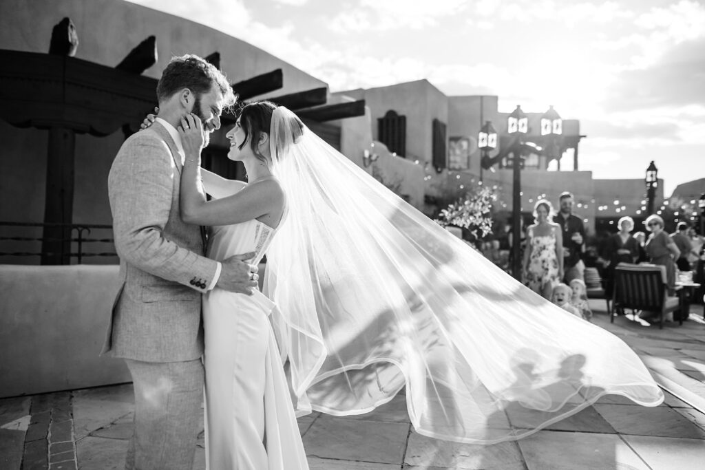 A black-and-white image of a bride and groom dancing on an outdoor terrace at sunset. The bride’s veil catches the wind as they embrace. Guests and string lights are visible in the background.