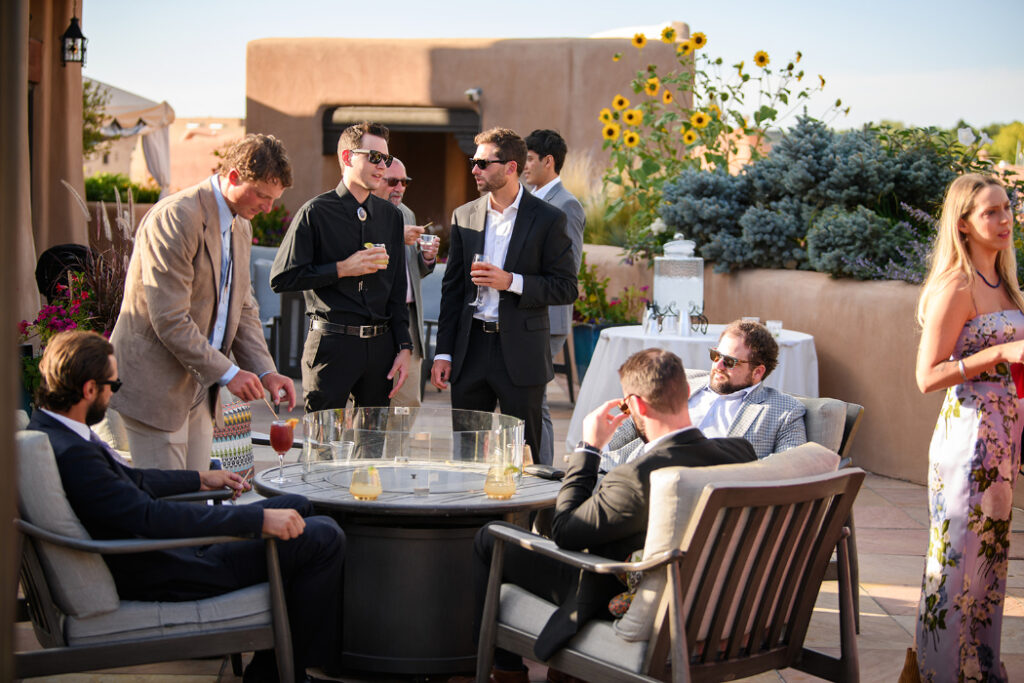 Wedding guests gather on an adobe-style rooftop patio for cocktail hour. Some sit around a fire pit, while others stand, holding drinks and chatting.