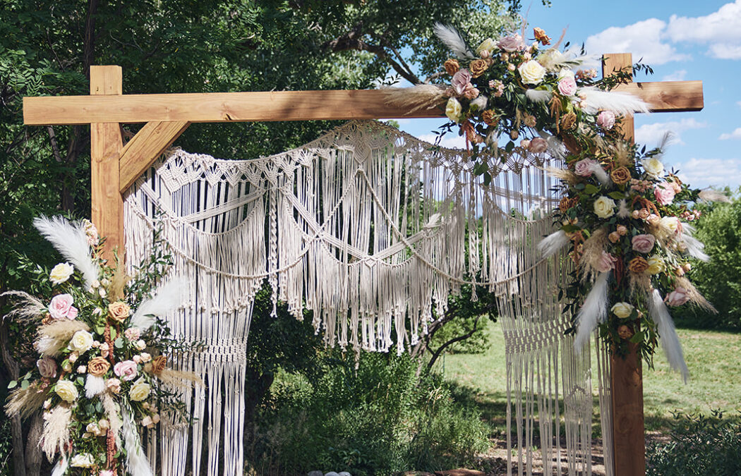 A wooden ceremony arch with macramé details and fresh floral arrangements stands against a backdrop of green trees and a bright blue sky.