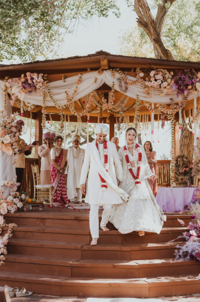 Bride and groom walk down steps smiling after their Indian wedding ceremony under a decorated mandap.