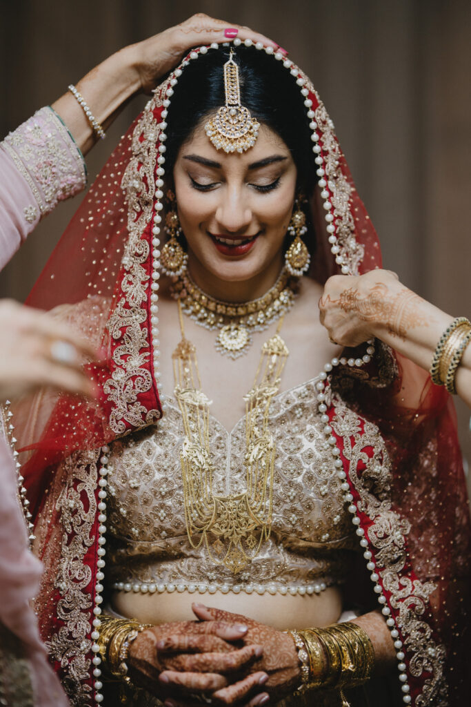 A bride in a red and gold lehenga has her dupatta carefully placed over her head. Her detailed jewelry, henna-adorned hands, and intricate embroidery highlight her traditional Indian bridal look.